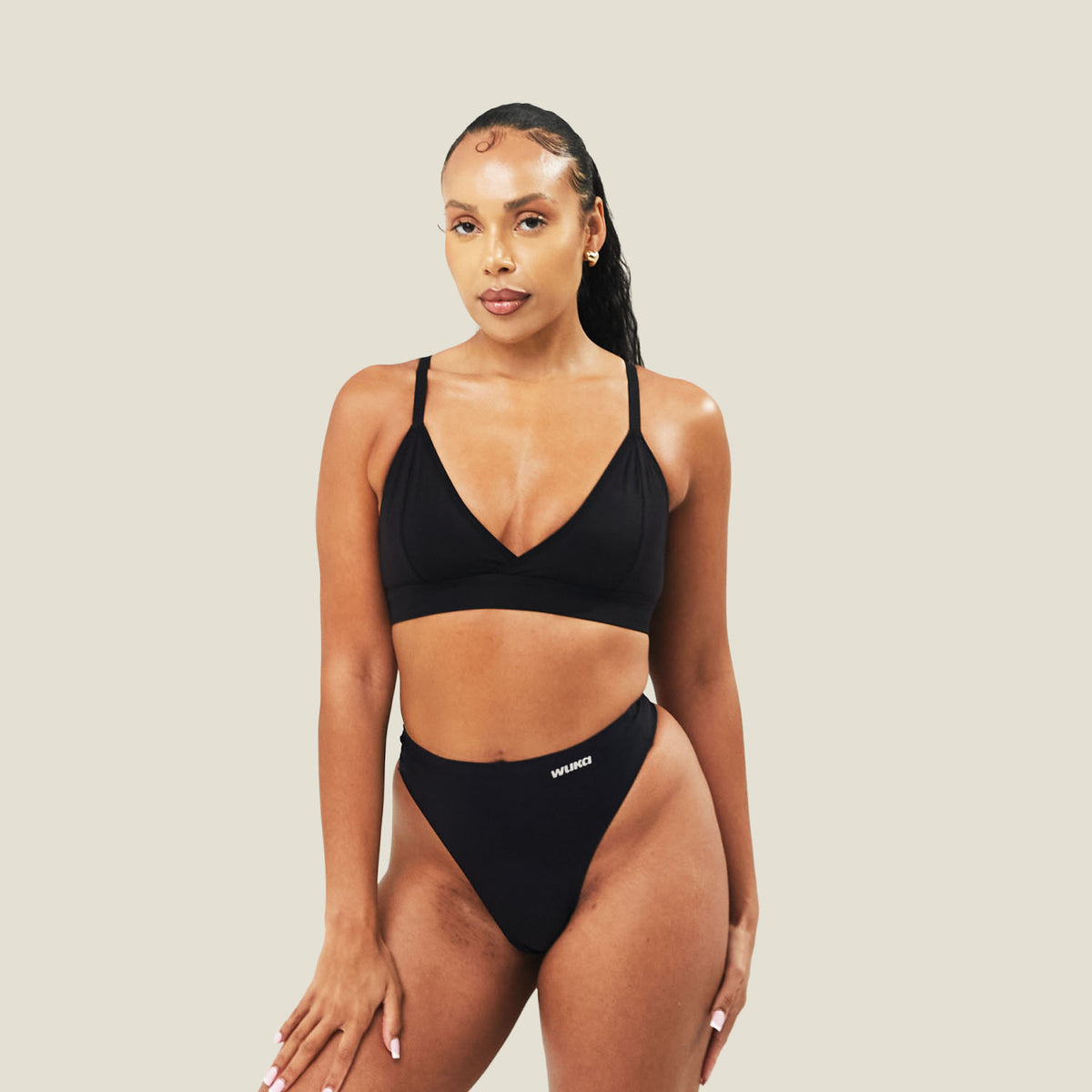 Model wearing the WUKA Stretch Seamless Mid Period Thong for light flow in black, with a matching black bralette, standing against a cream background.