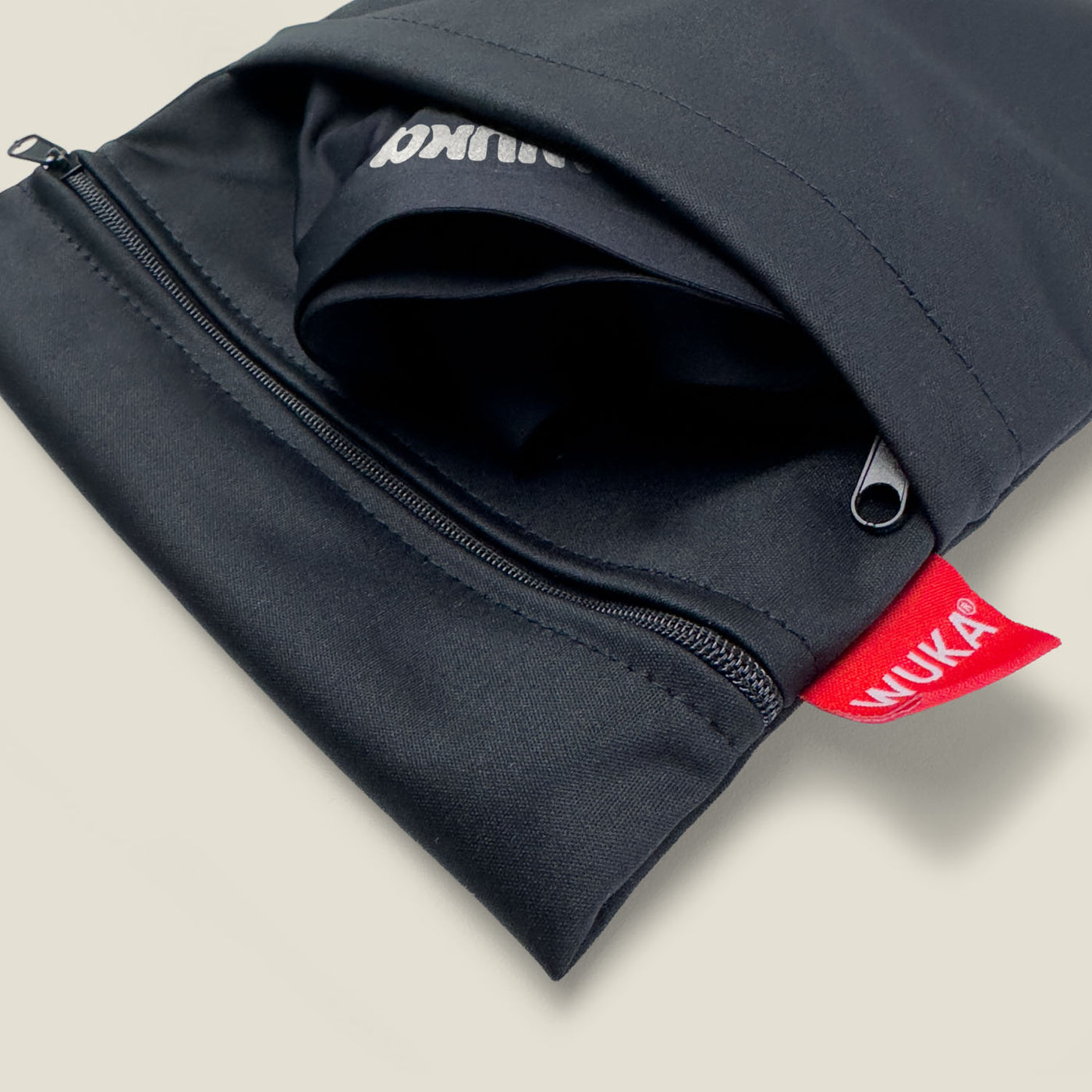 Close-up view of the WUKA Two Pocket Changing Bag showing zip detail and red branded tag, with folded WUKA pants partially tucked inside for convenient access.