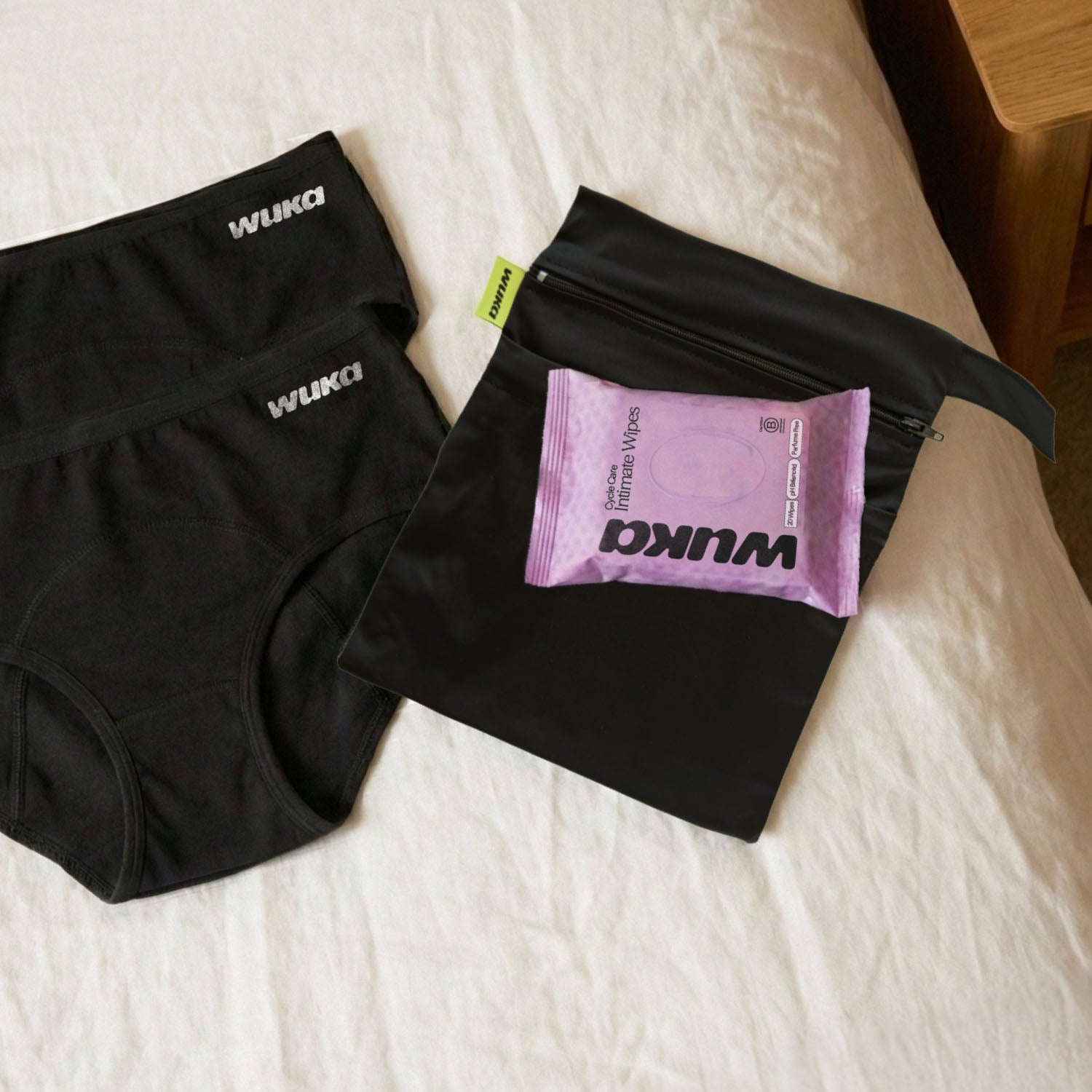 Lifestyle image of WUKA Go Bag laid on a bed, showing period pants, wipes, and 2-pocket changing bag in a real-world setting for everyday ease and comfort